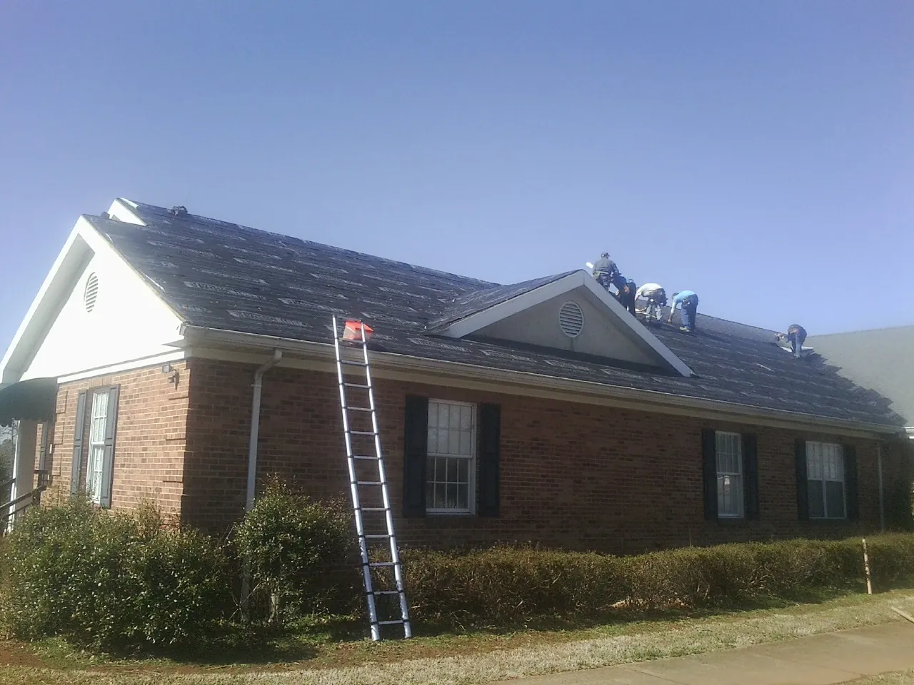 Crew tearing shingles off a brick ranch roof with a ladder set — Metal Roof Repair work by our crew in Orange Springs