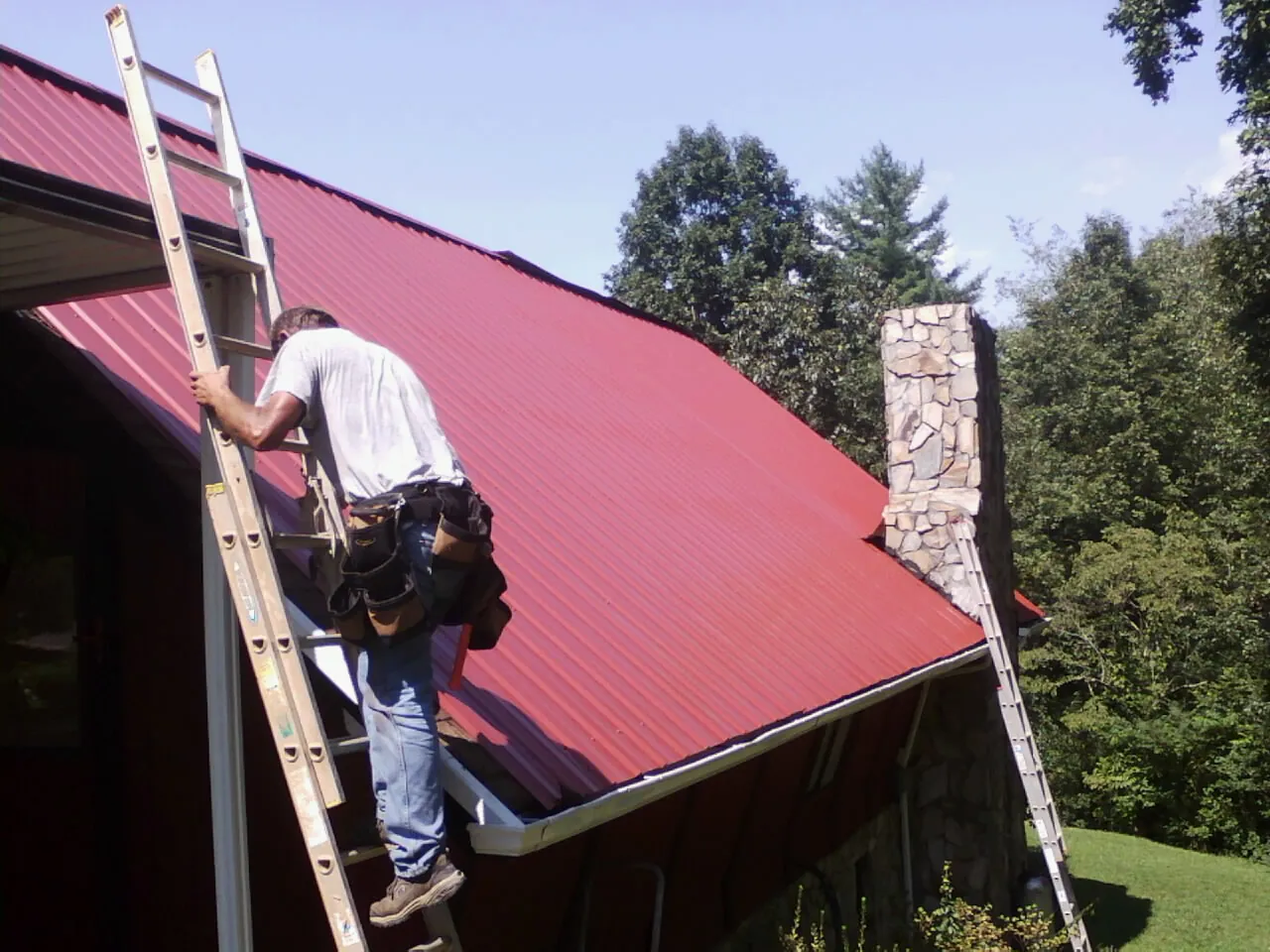 Roofer on a ladder beside a red metal roof and stone chimney — Soffit & Fascia Repair by Roofing Brothers in Orange Springs